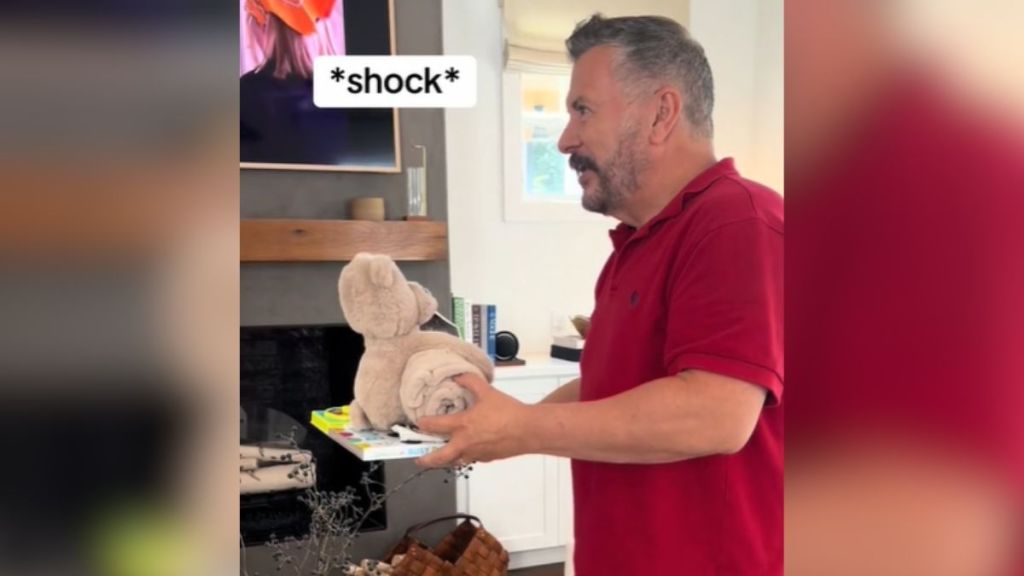 A man in a red shirt holding a teddy bear and looking surprised.