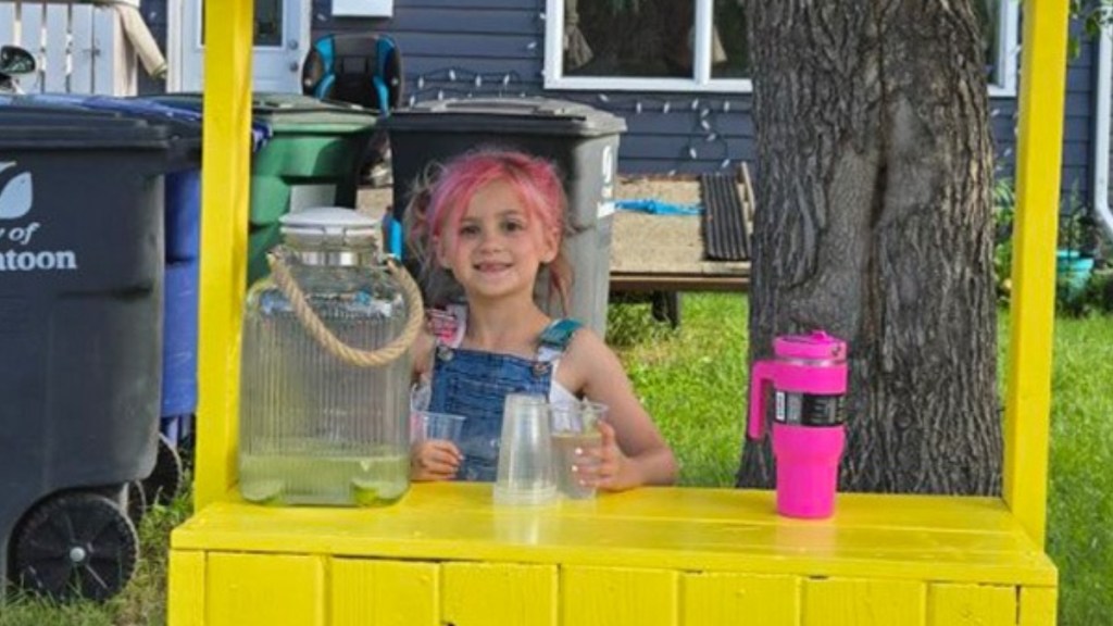 Close up of a little girl smiling from behind her lemonade stand. She's posing with a cup in hand. Next to her on the table is a glass container of lemonade.