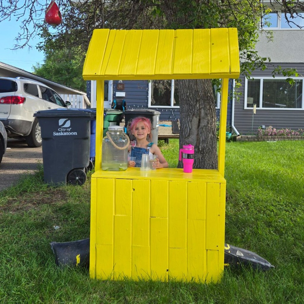 A little girl smiles from behind her lemonade stand. She's posing with a cup in hand. Next to her on the table is a glass container of lemonade. 