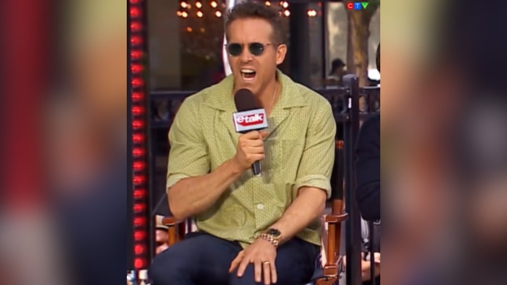 Ryan Reynolds looks shocked, mouth open, as he sits in a chair during an interview. He has a mic in hand and is wearing sunglasses