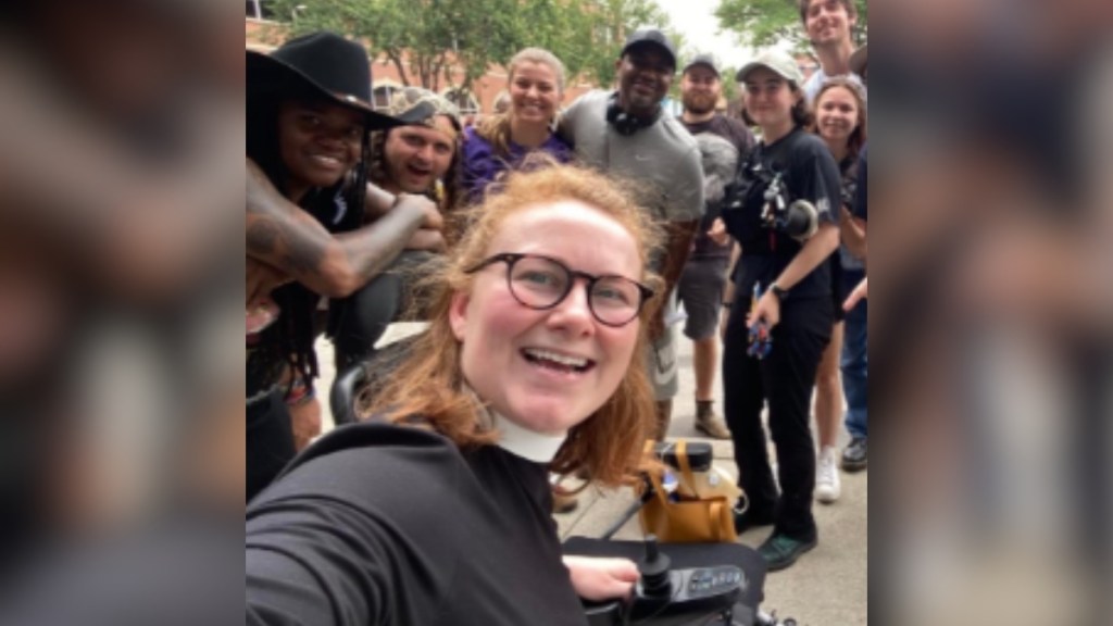 A woman smiles as she takes a selfie from her wheelchair. A whole group of people smiles and poses behind her