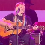 Willie Nelson smiles as he performs on stage. He sits as he plays guitar and sings