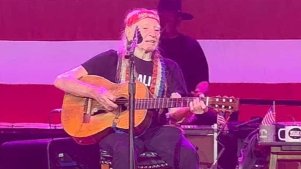 Willie Nelson smiles as he performs on stage. He sits as he plays guitar and sings