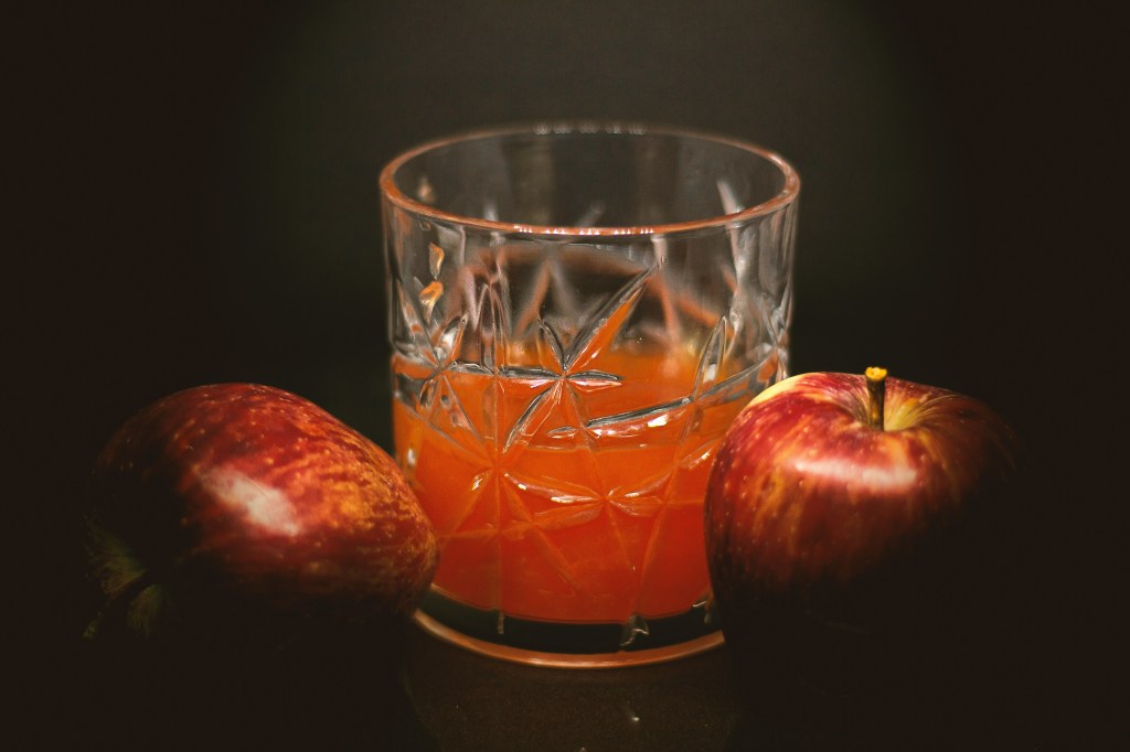 A small glass of apple juice. There are two apples next to the class.