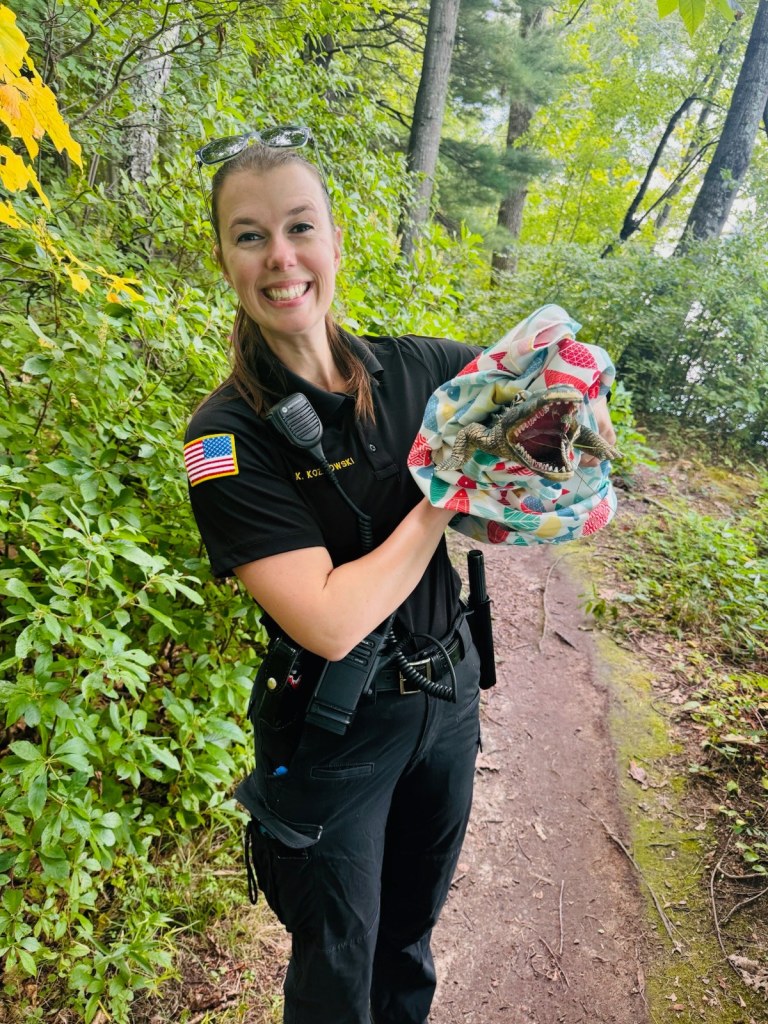 ACO Katie smiles wide as she holds up a small toy alligator wrapped in a blanket. The toy gator has its mouth wide open, showing off its teeth