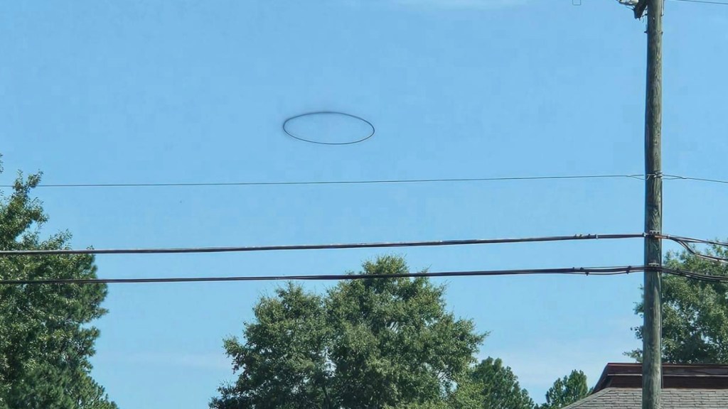 From a distance, a black ring floats in the bright blue sky above powerlines and trees