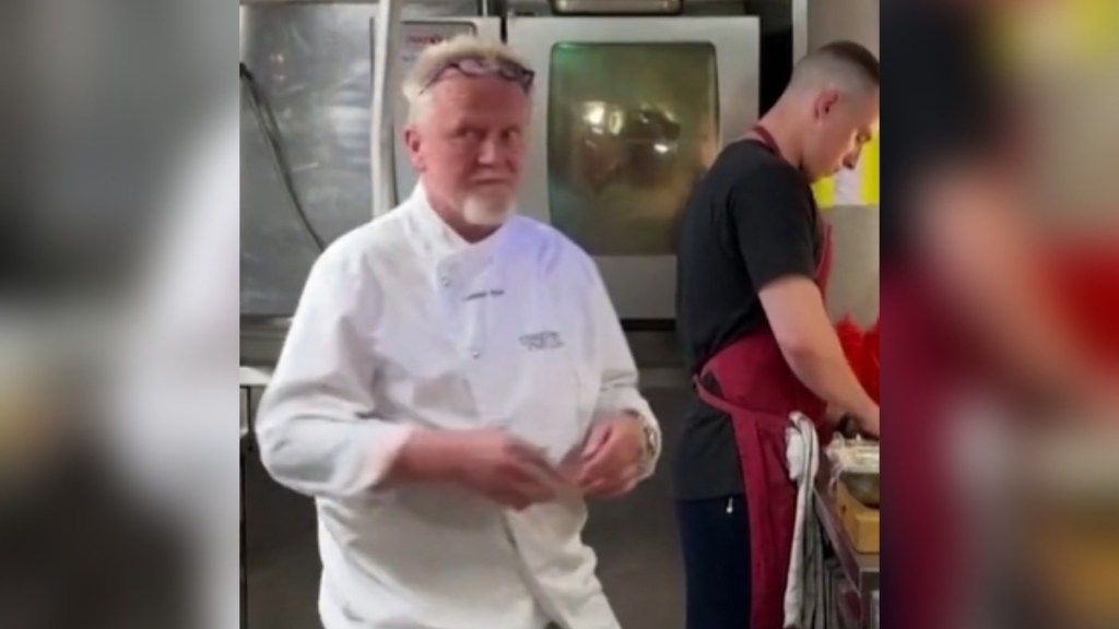 A chef makes his way through a kitchen, but we only see him from the waist up. A metal bar is just behind him. A second chef is at a counter behind him, working