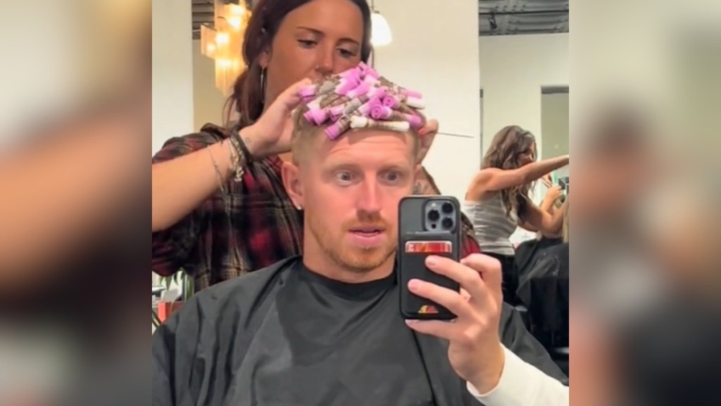 A man sits in a salon chair as a woman gives him a perm. There are curlers on his head. His eyes are wide, mouth slightly open, as he holds up his phone to capture the moment