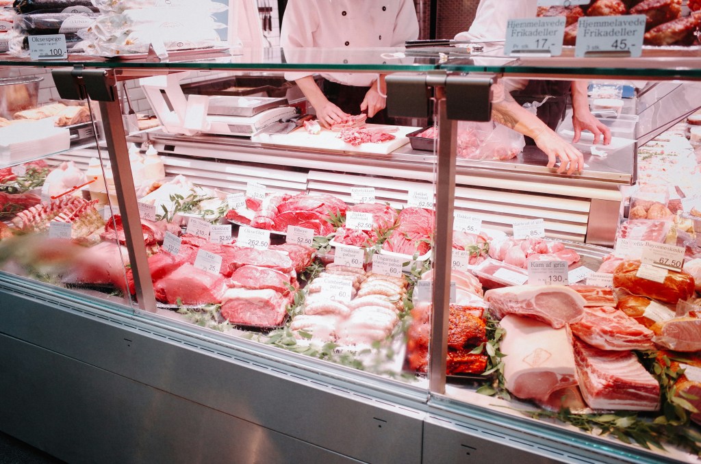 View of a deli with lots of meat options. Workers stand behind the counter, cutting and packaging meat