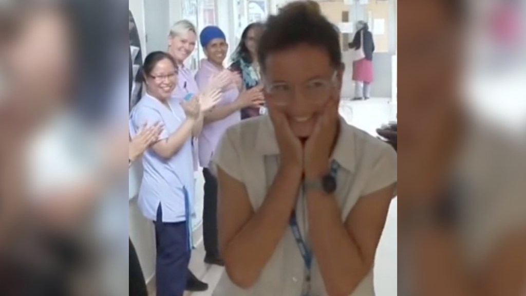 A woman smiles wide, hands on her face. She's walking down a hall where people are lined up, smiling and cheering for her