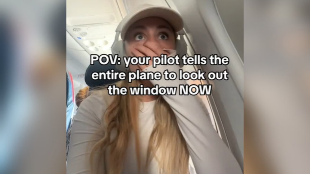 A woman sits on a plane, eyes wide as she covers her mouth with her hand. Text on the image reads: POV: your pilot tells the entire plane to look out the window NOW