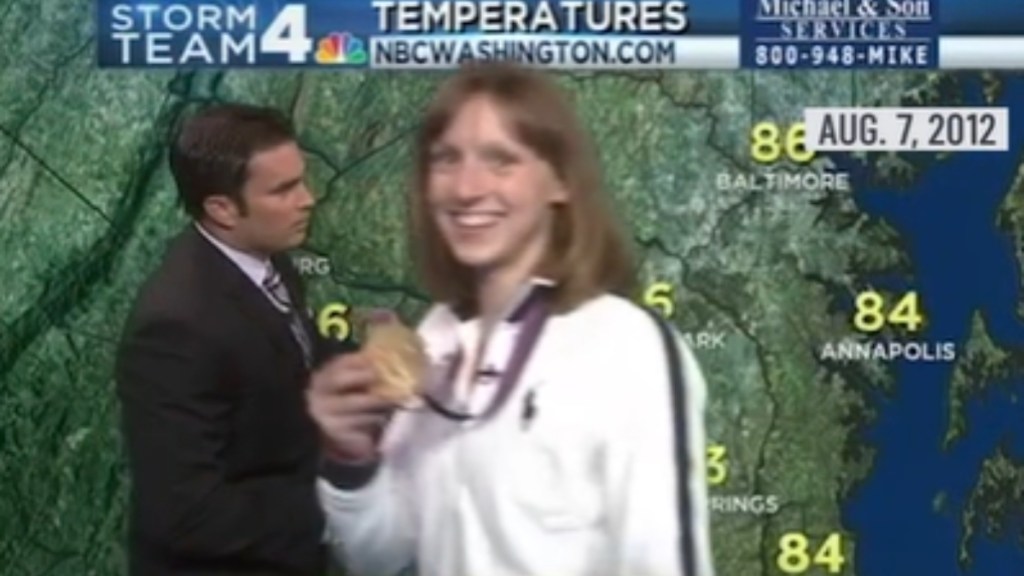 A teenage Katie Ledecky smiles as she walks in front of a meteorologist doing the news. She smiles as she holds up the gold medal around her neck