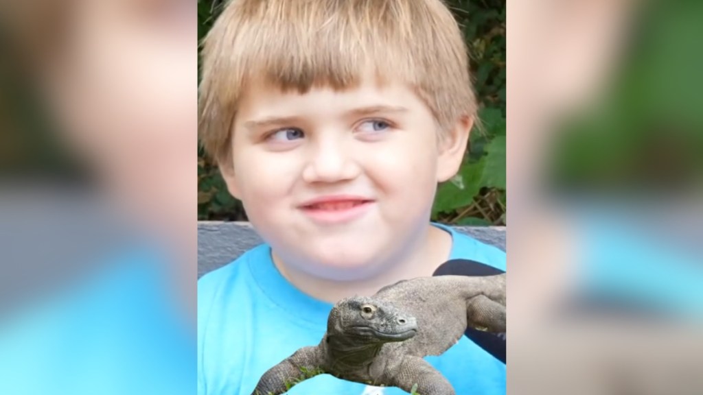 Close up of a little boy smiling as he looks off to the side. An image of a Komodo dragon has been edited onto the boy's chest/shoulder area