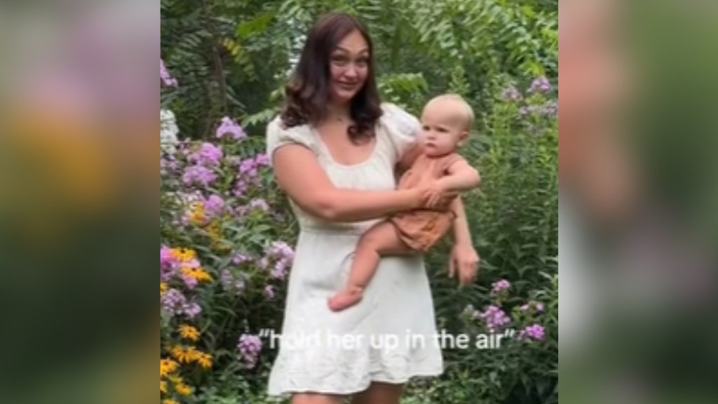 A woman stands outside with her baby in her arms. They stand in front of bushes with flowers. Mom's eyebrows are raised in surprise. Text on the image reads: "hold her up in the air"