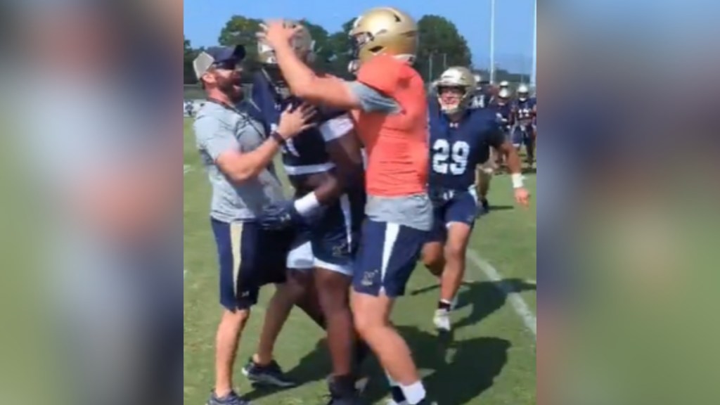 A football player is emotional as his coach and a fellow teammate surround him, excited. In the distance, other players can been seen approaching them