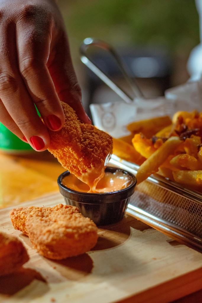 Close up of a chicken nugget being dipped into sauce. Other chicken nuggets are to the side. A pile of fries is on the other side