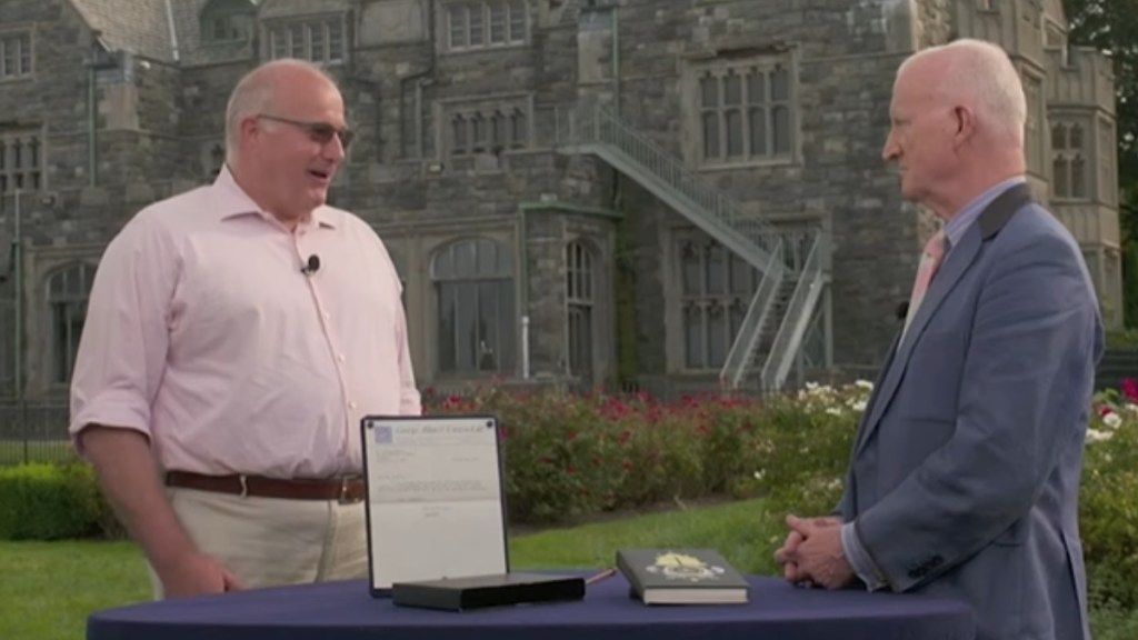 Two men stand outside at a table with a book and framed letter