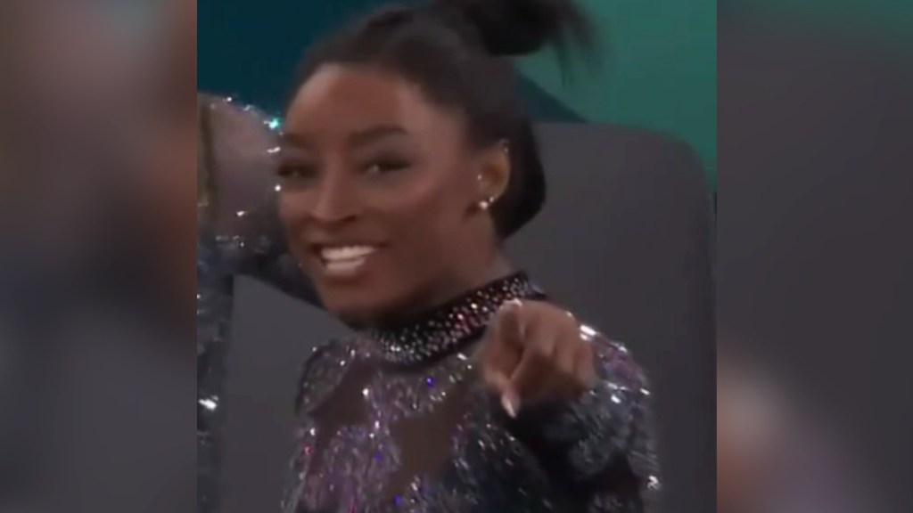 Simone Biles smiles wide as she points in the same direction as the camera.