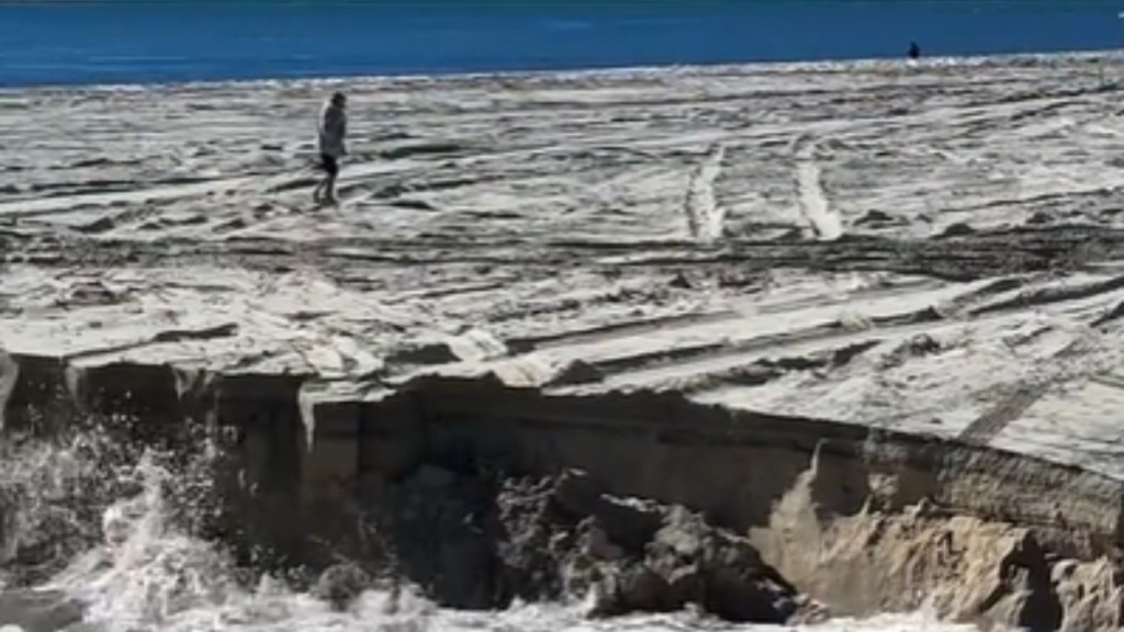 View of an Australian beach that's deteriorating from a sinkhole. Someone stands nearby at a distance, walking past