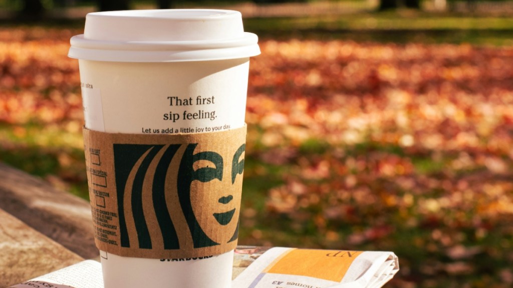 Close up of a Starbucks cup sat on a newspaper. In the background are fall leaves on the ground