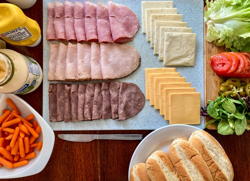 Top down view of a sandwich bar with deli meats, cheeses, lettuce, tomatoes, mayo, mustard, bread, and carrots