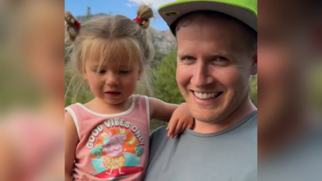 Close up of a dad smiling as he holds his grumpy toddler in his arms