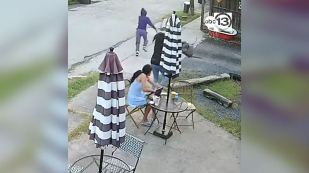 A woman steals a backpack from another woman sitting at a table.