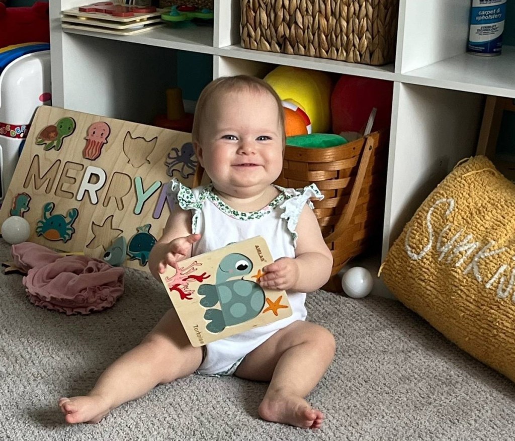 A baby smiles happily in front of an open, cubed storage unit with toys and other stuff. She's holding a baby book with a turtle on it