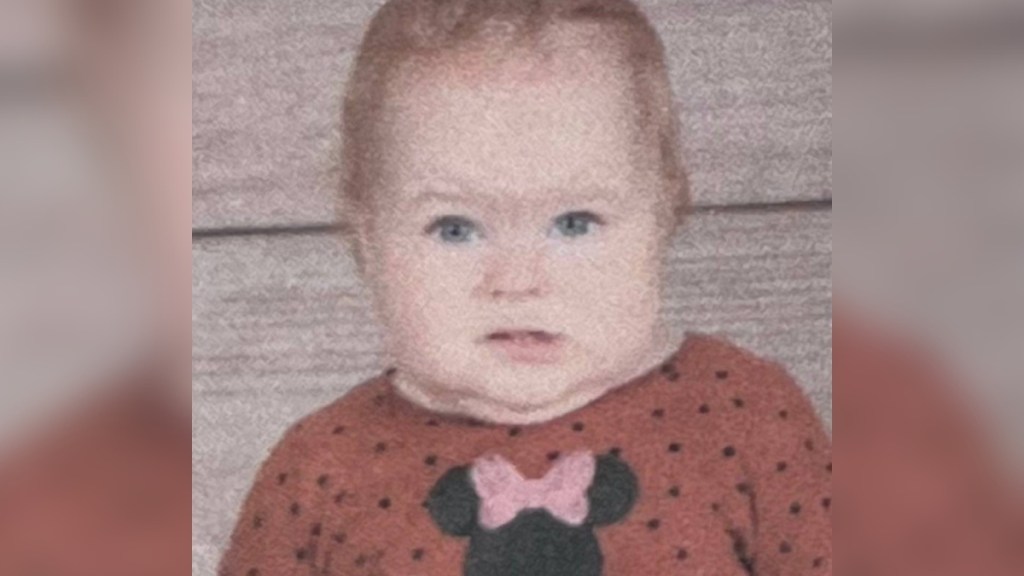 A baby gives an awkward smile, looking unsure of herself. A rag doll sits in her lap.