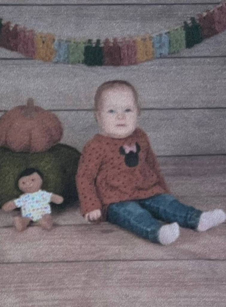 A grainy photo of a baby sitting in front of a natural backdrop. A colorful banner hangs on the wall. Next to the baby is a stack of two pumpkins. In front of the pumpkins is a small rag doll, arms splayed out by her sides with her legs splayed out in front of her. The baby, with a blank expression on her face, sits similarly to the doll