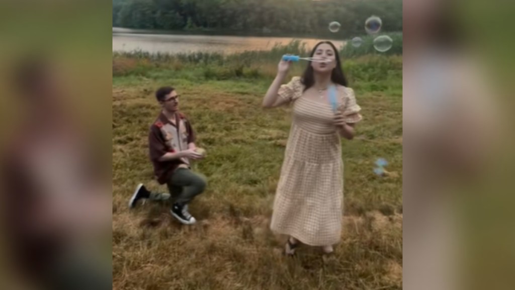 A man holds out a box with a ring while on one knee, ready to propose. A woman has her back away from him and faces the camera, happily blowing bubbles. They are in a beautiful field with water in the distance