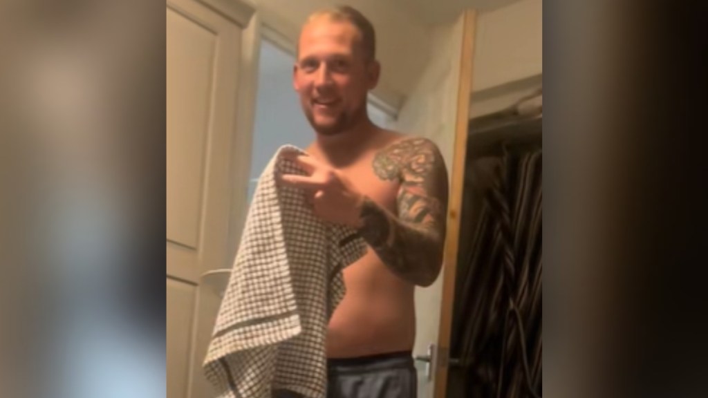 A man smiles wide as he carries something he's hiding behind a towel