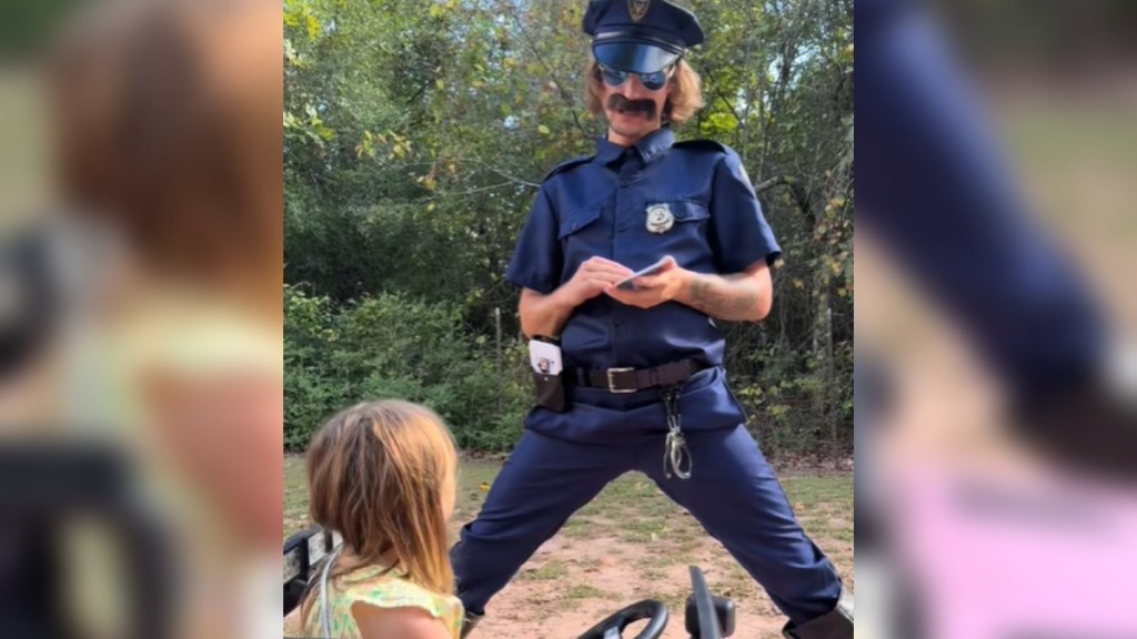 A man stands, legs spread wide, as he's dressed in a silly cop costume. He stands in front of a little girl who sits in a toy car, looking up at him