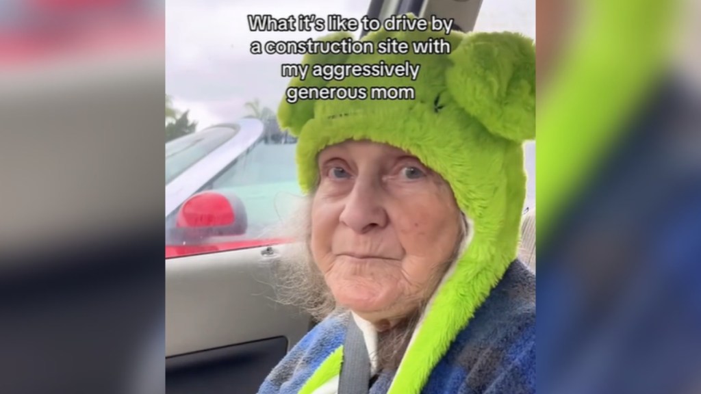 Close up of a 77 year old woman sitting in a car wearing a cute frog hat. She has a small smile on her face. Text on the image reads: What it's like to drive by a construction site with my aggressively generous mom