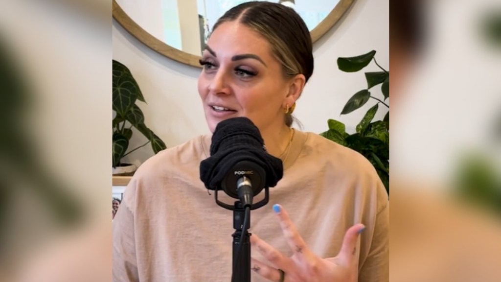 A woman uses her hands as she talks into a mic. She has a small smile on her face