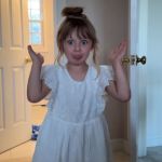 A little girl in a white dress making a silly face.