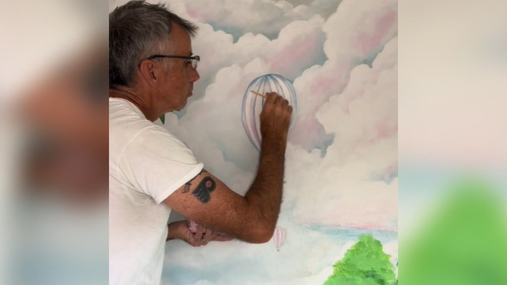 A man concentrates as he paints an air balloon on a mural amidst clouds and a nearby tree