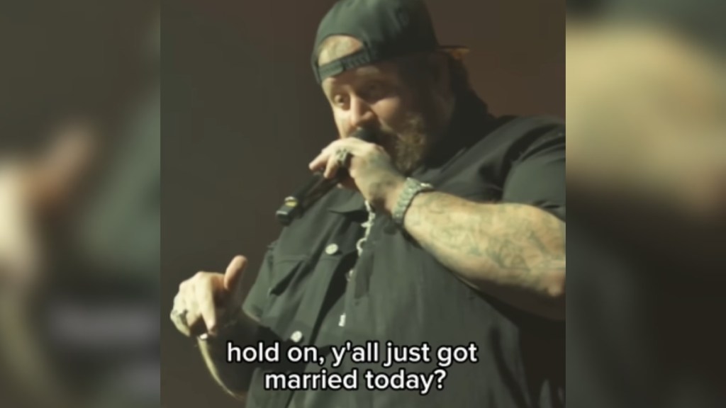 Close up of Jelly Roll talking into a mic. He points down to someone we can't see. His eyes are slightly wide, a look of surprise on his face. Text on the image reads: "Hold on, y'all just got married today?"