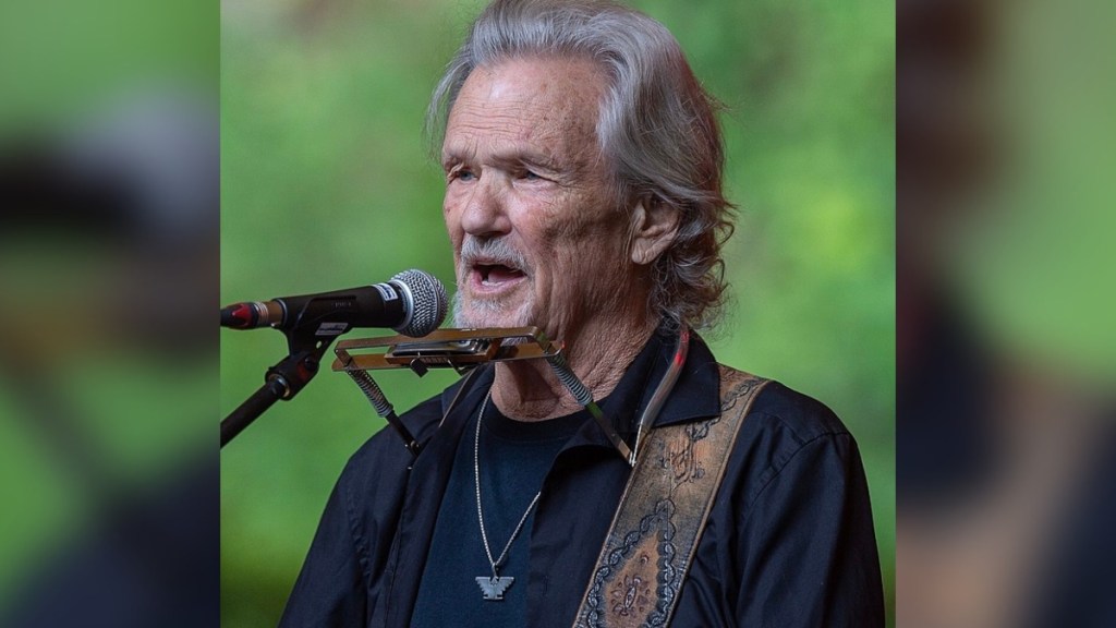 Close up of Kris Kristofferson performing. He sings into a mic while wearing a devise that allows him to play a harmonica hands free