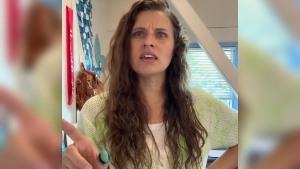 Close up of a teacher as she talks. One hand is on her hip while she uses the other to point