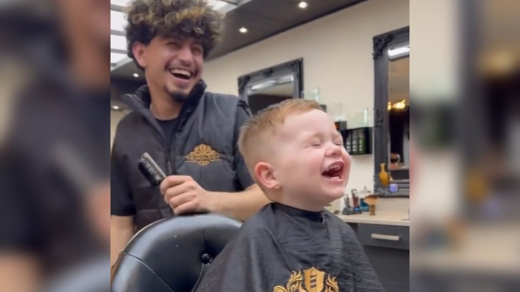 A little boy sits in a barber chair, laughing so hard his eyes are closed and his mouth is wide open. The barber stands behind him, laughing just as hard