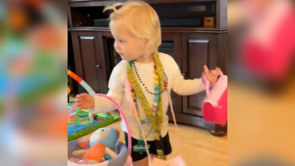 A toddler looks off to the side as she walks around with her arms full of purses and a doll. She's also wearing a ton of necklaces