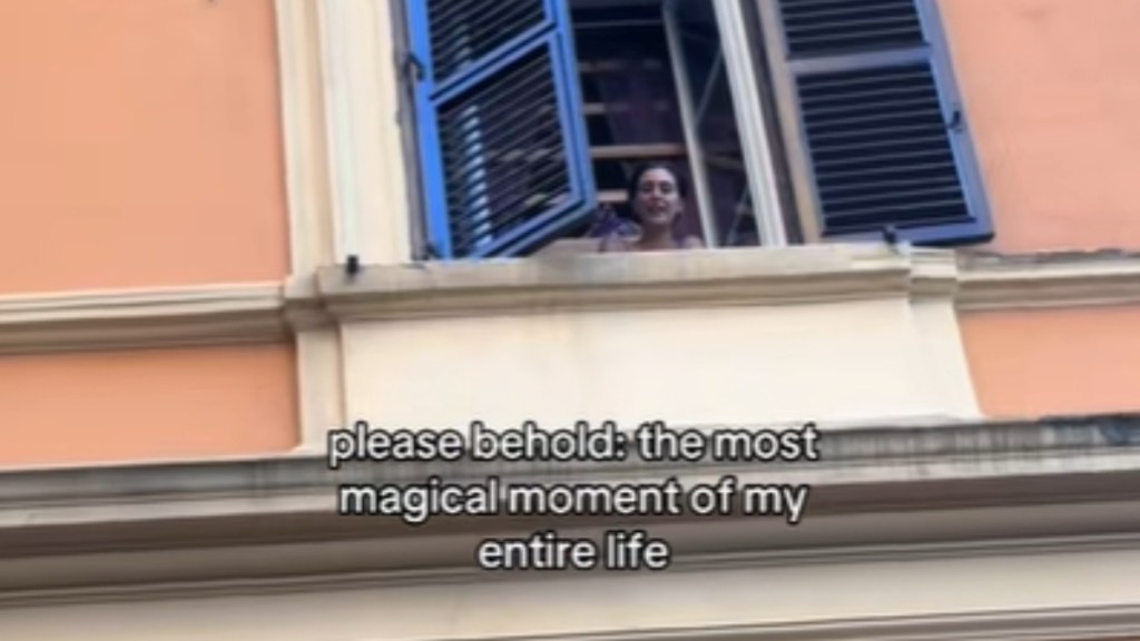 Close up of a woman who is at a distance, peaking her head out of her apartment window. She smiles, mouth open. Text on the image reads: please behold the most magical moment of my entire life