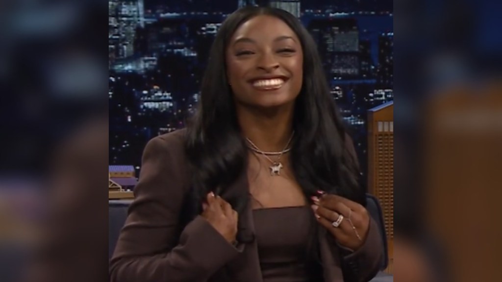 Simone Biles smiles wide as she sits during an interview with Jimmy Fallon. She moves back her hair to showoff the goat necklace she's wearing