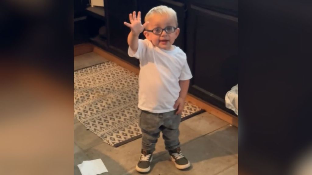 A toddler with glasses holding up one hand.