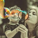 A woman holds a little boy in her arms as she blows giant bubbles. The boy smiles as he watches