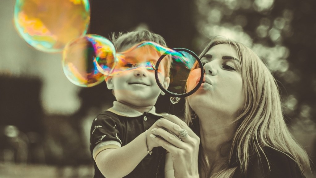 A woman holds a little boy in her arms as she blows giant bubbles. The boy smiles as he watches