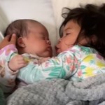 Close up of a baby and a toddler snuggled under a blanket in a large bed. The toddler seems to be moving her mouth, one arm around the front of the baby. The baby has one hand gently placed on the toddler's arm. The baby's eyes are closed