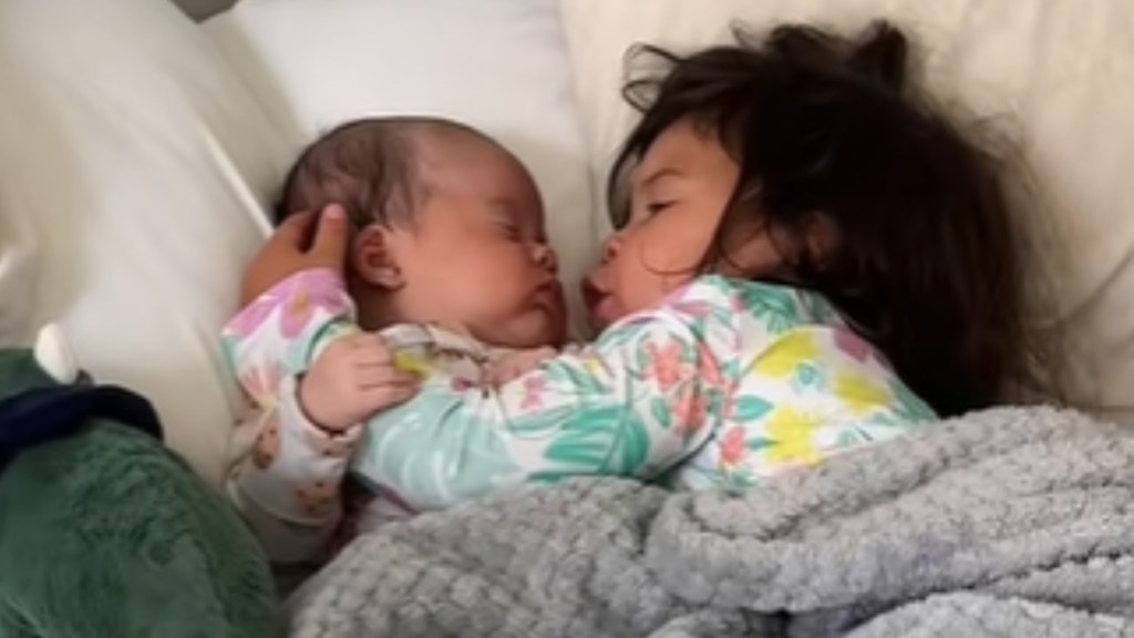 Close up of a baby and a toddler snuggled under a blanket in a large bed. The toddler seems to be moving her mouth, one arm around the front of the baby. The baby has one hand gently placed on the toddler's arm. The baby's eyes are closed