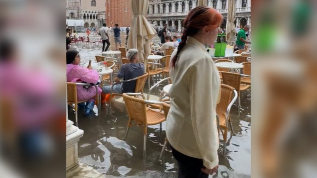 A waiter walks past the camera with a tray. Nearby, patrons sit at tables in a courtyard. The flooding is so high that they're all wearing rain boots